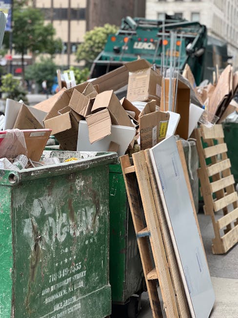 A large green waste skip overflowing with various types of rubbish including cardboard boxes, wooden pallets, and packaging materials, positioned on a street in an urban environment. In the background, a commercial garbage collection truck is partially visible, indicating ongoing waste collection activity. The scene is outdoors with city buildings, trees, and parked vehicles surrounding the area. Visible environmental details include the pavement and nearby greenery. The materials in the skip consist of corrugated cardboard, wooden planks, and white packaging boxes, some of which are crushed or jumbled. The overall scene reflects a private or alternative waste handling scenario outside of standard council rubbish collection, emphasizing the importance of professional rubbish removal services like those offered by Waste Disposal Docklands in managing bulky or mixed waste effectively in congested or narrow access locations. The lighting suggests natural daylight, providing clear visibility of the waste stored in the skip and the surrounding urban setting.