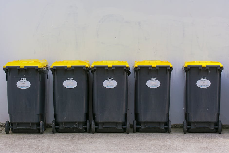 A row of five black wheeled rubbish bins with yellow lids, aligned along a light grey concrete surface and positioned against a plain, light-colored wall. Each bin has a white barcode label affixed to the front, with some labels displaying numbers such as 35110, 35181, 35122, 35207, and 35297. The lids are securely closed, and the bins are evenly spaced, showing slight variations in wear and dirt marks on their surfaces. The environment appears to be an outdoor area possibly adjacent to a commercial or residential building, suitable for private waste collection or alternative rubbish disposal services. The plain background and uniformity of the bins emphasize their primary function of waste storage and collection, making the scene relevant to waste management operations such as private rubbish collection or on-site refuse handling, facilitated by services like Waste Disposal Docklands.