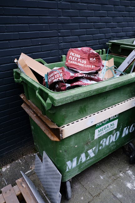 A large green waste skip overflowing with various types of rubbish including cardboard boxes, wooden pallets, and packaging materials, positioned on a street in an urban environment. In the background, a commercial garbage collection truck is partially visible, indicating ongoing waste collection activity. The scene is outdoors with city buildings, trees, and parked vehicles surrounding the area. Visible environmental details include the pavement and nearby greenery. The materials in the skip consist of corrugated cardboard, wooden planks, and white packaging boxes, some of which are crushed or jumbled. The overall scene reflects a private or alternative waste handling scenario outside of standard council rubbish collection, emphasizing the importance of professional rubbish removal services like those offered by Waste Disposal Docklands in managing bulky or mixed waste effectively in congested or narrow access locations. The lighting suggests natural daylight, providing clear visibility of the waste stored in the skip and the surrounding urban setting.