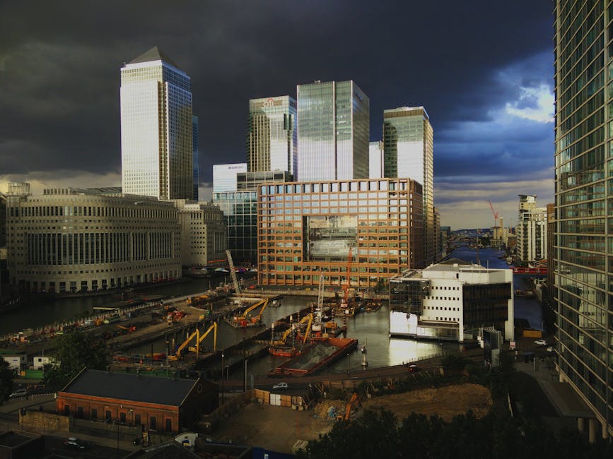 A wide view of a construction site situated in a modern urban area, with several yellow cranes actively lifting and moving materials near the water's edge. The site features large, rectangular construction vessels and heavy machinery, with piles of construction materials and equipment visible on the quay. Surrounding the site are tall office buildings with reflective glass facades and a distinctive orange-brown brick building with a large rectangular opening in the middle, which appears to be part of the ongoing development. The background includes dark, stormy clouds contrasting with patches of blue sky and sunlight illuminating the upper stories of the surrounding skyscrapers. This scene exemplifies a typical active construction zone within a financial district, highlighting the importance of effective waste and rubbish management, as provided by services such as Waste Disposal Docklands, during periods of large-scale urban development or on-site clearance activities.