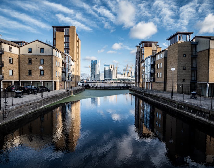 A canal lined with modern, multi-story residential buildings constructed from tan and beige bricks, with black-framed windows and small balconies on the upper floors. The buildings are situated on both sides of the water, with paved walkways and black metal railings along the canal edge. Several street lamps are evenly spaced along the walkway, and a row of parked cars is visible to the left. In the background, a bridge spans the canal, leading to a cluster of contemporary office buildings and a large construction crane, indicating ongoing development. The sky above features scattered clouds with patches of blue, and the water reflects the facades of the buildings and the sky, creating a calm, mirrored surface, emphasizing the urban environment and clean setting of the area managed by Waste Disposal Docklands for private waste removal and on-site clearance services.