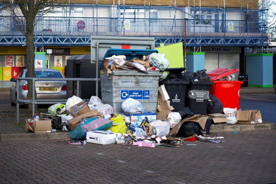 A large pile of mixed domestic waste and rubbish, including cardboard boxes, plastic bags, paper debris, and discarded packaging, sits overflowing from a metal recycling bin with a blue lid and a label indicating it is for mixed paper and cardboard. Surrounding the bin, additional black and red rubbish bags, flattened boxes, and loose paper are scattered on the paved area of a parking lot. Behind the rubbish, there is a parked silver car with a yellow license plate, and a metal railing separates the waste area from the vehicle. In the background, a retail unit with storefronts, signage, and a blue scaffolding structure suggests an urban commercial environment. The scene is illuminated by natural daylight, highlighting the cluttered, unmanaged appearance of the waste, which is indicative of off-site rubbish collection practices handled by waste disposal services such as Waste Disposal Docklands.