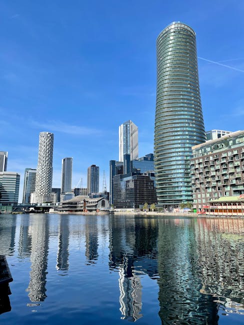 A canal lined with modern, multi-story residential buildings constructed from tan and beige bricks, with black-framed windows and small balconies on the upper floors. The buildings are situated on both sides of the water, with paved walkways and black metal railings along the canal edge. Several street lamps are evenly spaced along the walkway, and a row of parked cars is visible to the left. In the background, a bridge spans the canal, leading to a cluster of contemporary office buildings and a large construction crane, indicating ongoing development. The sky above features scattered clouds with patches of blue, and the water reflects the facades of the buildings and the sky, creating a calm, mirrored surface, emphasizing the urban environment and clean setting of the area managed by Waste Disposal Docklands for private waste removal and on-site clearance services.