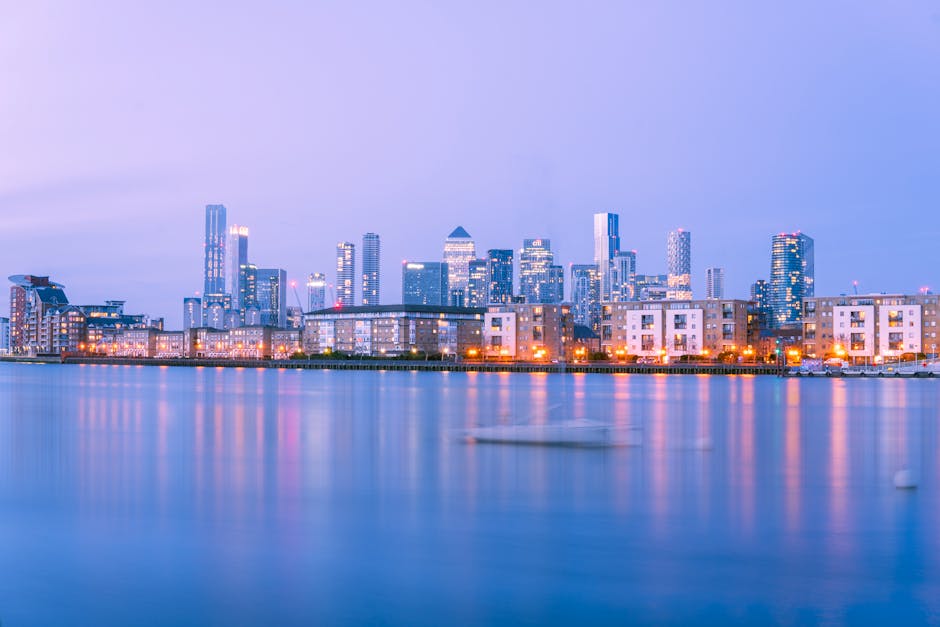 A panoramic view of a modern city skyline at dusk, featuring numerous high-rise office buildings and residential towers illuminated with warm and cool lighting, situated along a waterfront with a calm body of water reflecting the lit structures. In the foreground, a paved area with a low boundary wall is visible, with some buildings appearing to be low-rise apartment blocks or commercial units. The sky is overcast with a soft, muted hue, and the overall scene suggests an urban environment near a river or harbor, aligning with the context of waste disposal and potential for on-site clearance or alternative rubbish collection services provided by Waste Disposal Docklands. The multiple buildings’ designs range from glass facades to concrete finishes, and there are faint silhouettes of boats on the water, adding to the environmental setting. The lighting highlights the contrast between the bustling city and the tranquil water, reflecting a typical cityscape scene suitable for illustrating rubbish removal or waste management services in an urban environment near a waterway.