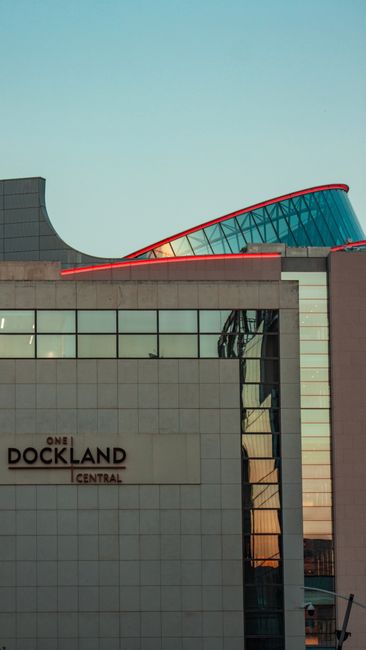 The image shows the upper section of a modern building with a curved, glass-covered rooftop structure featuring red framing, set against a clear blue sky during daytime. The lower part of the building is clad in light-colored, smooth tiles with reflective windows, one of which reveals a glimpse of the interior and structural elements like steel supports. A sign reading 'ONE DOCKLAND CENTRAL' is visible on the facade, indicating a commercial or office property in Docklands. The overall scene is well-lit, with natural daylight highlighting the architectural details and the sleek, contemporary design of the building. This setting suggests an urban environment within the Docklands area, where high-rise commercial developments often require regular rubbish clearance services, such as those provided by Waste Disposal Docklands, to manage waste from offices and retail outlets efficiently.