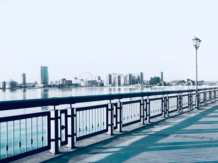 A panoramic view of a modern city skyline at dusk, featuring numerous high-rise office buildings and residential towers illuminated with warm and cool lighting, situated along a waterfront with a calm body of water reflecting the lit structures. In the foreground, a paved area with a low boundary wall is visible, with some buildings appearing to be low-rise apartment blocks or commercial units. The sky is overcast with a soft, muted hue, and the overall scene suggests an urban environment near a river or harbor, aligning with the context of waste disposal and potential for on-site clearance or alternative rubbish collection services provided by Waste Disposal Docklands. The multiple buildings’ designs range from glass facades to concrete finishes, and there are faint silhouettes of boats on the water, adding to the environmental setting. The lighting highlights the contrast between the bustling city and the tranquil water, reflecting a typical cityscape scene suitable for illustrating rubbish removal or waste management services in an urban environment near a waterway.