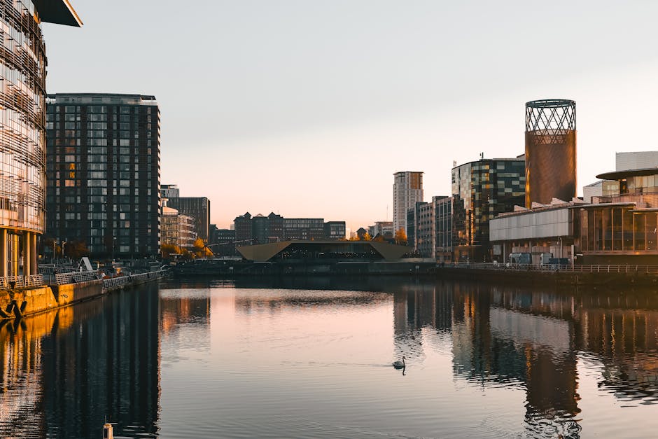 A panoramic view of a modern urban waterfront during sunset, featuring tall commercial and residential buildings with glass facades reflecting the soft orange and pink hues of the sky. The buildings are positioned on either side of a calm river or canal, with their mirrored images visible in the water. On the left, a rounded high-rise with a glass exterior shows vertical and horizontal window patterns, while on the right, a distinctive cylindrical tower with a lattice-like top section is prominent. The water surface is smooth, with slight ripples, and a solitary bird can be seen floating near the center. The scene suggests a clean, well-maintained environment, typical of a city area where private waste management companies, such as Waste Disposal Docklands, may assist with rubbish removal or on-site clearance, especially in settings with modern infrastructure and waterway access. The overall atmosphere is tranquil, emphasizing the neatness and urban planning of the area consistent with local council rules for waste management and bulky rubbish collection.