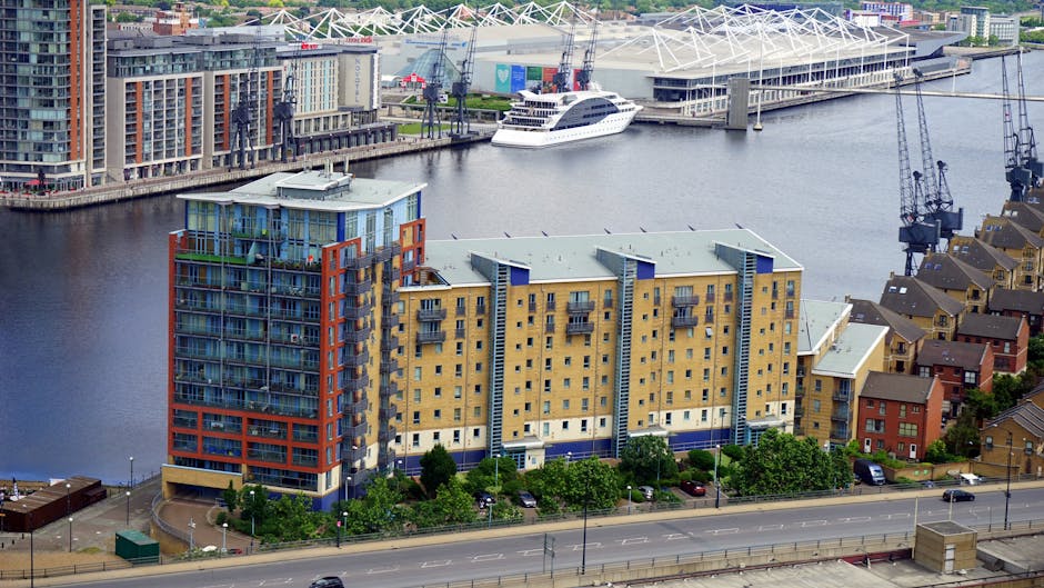 An aerial view of a multi-storey residential building with a modern design, featuring a combination of beige and blue exterior walls, numerous small balconies with glass railings, and several trees and parked cars lining the street below. In the background, a body of water hosts a large white yacht docked near a waterfront area with construction cranes and a sports stadium with a distinctive white roof structure. The scene captures the urban environment typical of the Docklands area, illustrating a mix of residential, commercial, and recreational spaces, and reflecting the context of private waste disposal and alternative rubbish collection methods. The lighting suggests a clear day with natural daylight illuminating the scene evenly, providing a neutral and detailed representation of the environment.