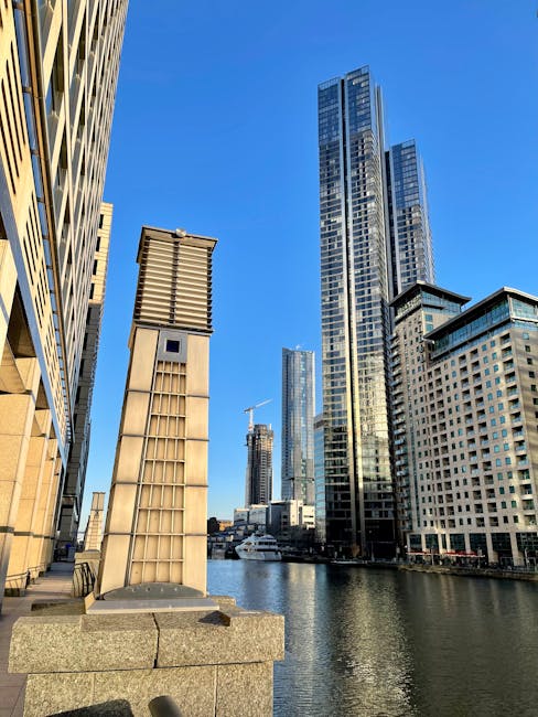 A panoramic view of a modern urban waterfront during sunset, featuring tall commercial and residential buildings with glass facades reflecting the soft orange and pink hues of the sky. The buildings are positioned on either side of a calm river or canal, with their mirrored images visible in the water. On the left, a rounded high-rise with a glass exterior shows vertical and horizontal window patterns, while on the right, a distinctive cylindrical tower with a lattice-like top section is prominent. The water surface is smooth, with slight ripples, and a solitary bird can be seen floating near the center. The scene suggests a clean, well-maintained environment, typical of a city area where private waste management companies, such as Waste Disposal Docklands, may assist with rubbish removal or on-site clearance, especially in settings with modern infrastructure and waterway access. The overall atmosphere is tranquil, emphasizing the neatness and urban planning of the area consistent with local council rules for waste management and bulky rubbish collection.