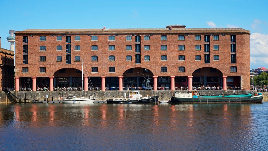 A large red brick building situated along a riverbank, with four pink-painted columns supporting the ground floor structure and creating open archways. The building has a flat roof and multiple small, uniformly spaced windows on its upper floors, with some windows having black frames and grid-like panes. Docked directly in front of the building are three boats: a white sailboat with a mast on the left, a small white motorboat in the middle, and a longer, dark green barge on the right. The boats are moored to a concrete quay or wharf, which has a textured, weathered surface. The scene is illuminated by natural daylight, with a clear blue sky featuring a few scattered clouds in the background. This setting suggests a waterfront location with infrastructure suitable for maritime activities, where private or alternative waste disposal methods may operate outside conventional public rubbish collection services, aligning with the concept of independent rubbish removal often associated with professional waste management services like Waste Disposal Docklands.